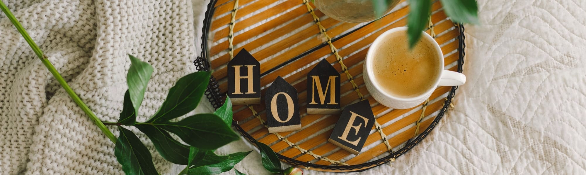 Un plateau en bambou avec une couverture tricot blanc, des feuille et un café avec 4 maisons bois déco formant HOME.